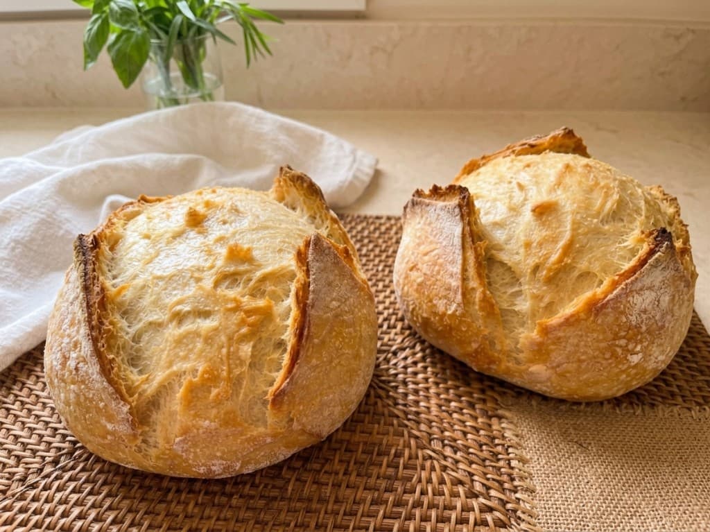 Two round artisan sourdough loaves on a woven mat with a linen cloth and herbs in the background